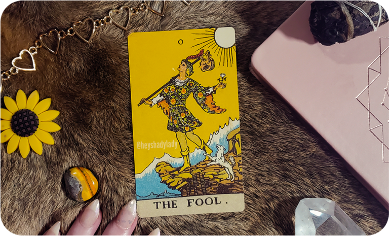an aesthetic photograph of The Fool tarot card in center frame with various items scattered around: a soft pink book with a golden geometric design in the center, a burned bundle of black sage, a clear quartz crystal, a bumblebee jasper mineral, a gold chain heart-shaped necklace, and a yellow sunflower decoration. all of these objects rest on a background of rabbit fur, and there are a few fingers with long french tip nails in the bottom of the frame.