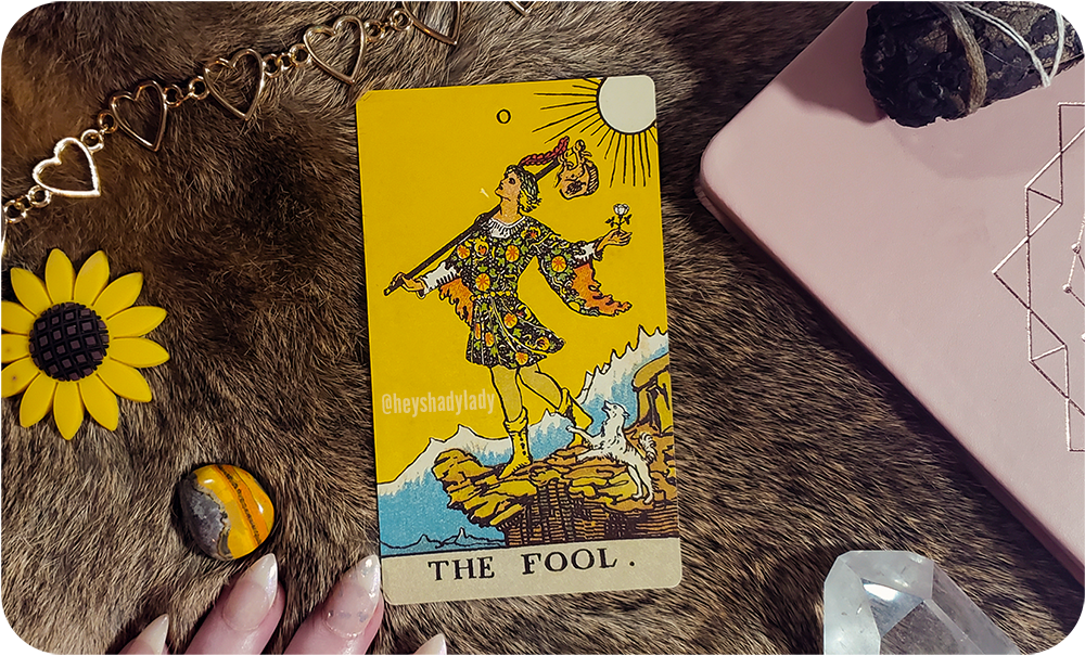 an aesthetic photograph of The Fool tarot card in center frame with various items scattered around: a soft pink book with a golden geometric design in the center, a burned bundle of black sage, a clear quartz crystal, a bumblebee jasper mineral, a gold chain heart-shaped necklace, and a yellow sunflower decoration. all of these objects rest on a background of rabbit fur, and there are a few fingers with long french tip nails in the bottom of the frame.