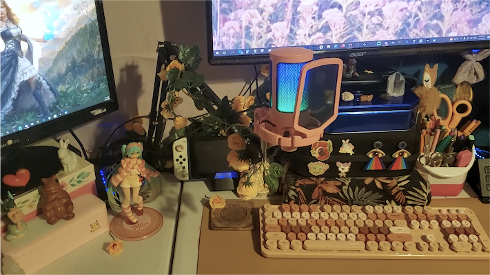 a very decorated and cozy desk setup, full of earthy and pastel colors.  you can see two monitors powered on with flowery fantasy wallpapers.  a full pen holder on the right next to a stream deck, crystals and various animal figurines decorating all surfaces of the deck. there's a nintendo switch and anime figurine of miku in the background. an aesthetic, old-fashioned looking keyboard is in the foreground, but the center piece is the plugged in, assembled and lit-up fifine microphone, now adorned with fake peachy-colored flowers as decoration.