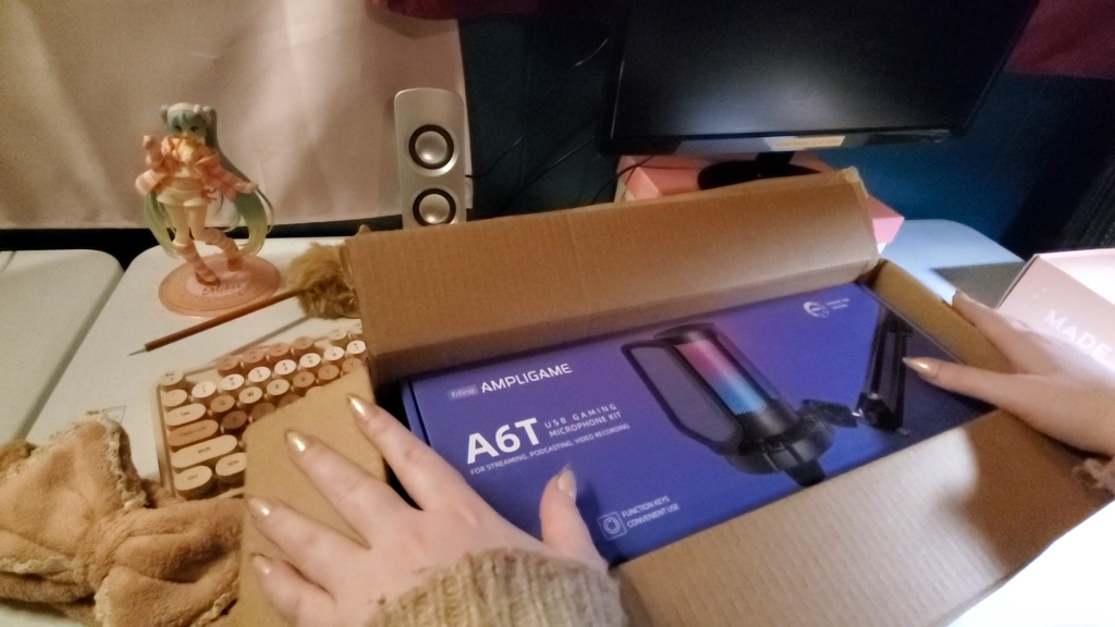 a picture of a bare desk setup, one monitor and speaker are visible in the background. a cute anime figurine of miku from vocaloid is positioned in the back.  there is an open package in the middle of the desk with a purple box visible.  there is a microphone on the cover of the box with the label "ampligame A6T usb caming microphone kit." heyshadylady's hands are resting on either side of the box.