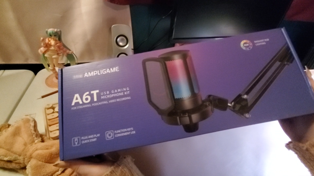 a picture of a bare desk setup, one monitor and speaker are visible in the background. a cute anime figurine of miku from vocaloid is positioned in the back.  heyshadylady is using both hands to hold up an unopened purple box that has a microphone on the cover and a label reading "ampligame A5T usb gaming microphone kit"