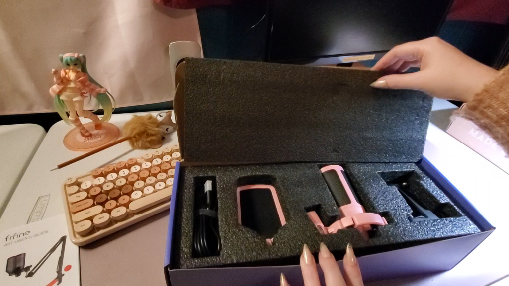 a picture of a bare desk setup, one monitor and speaker are visible in the background. a cute anime figurine of miku from vocaloid is positioned in the back, along with a cute pen with a fuzzy fox decorating the top, and an aesthetic keyboard.  there is a user manual in the corner of the picture for the fifine mirophone, and heyshadylady is peeling back a black styrofoam lid to reveal four parts of the unassembled microphone: a USB cord, a pop filter, the microphone head and the metal piece used to attach the microphone's boom arm to the desk. all of these pieces are nestled deep into a black protective styrofoam container.