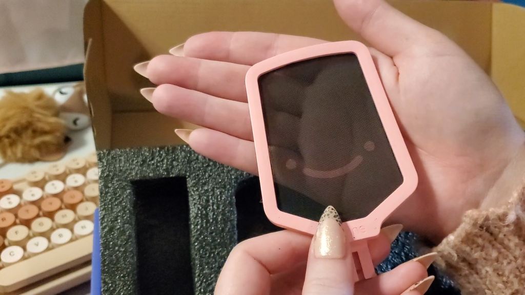 a close up photo of heyshadylady's hands holding the fifine microphone pop filter.  the pop filter is a tall rectangular shape with a soft pink border, and solid black screen. the filter screen has an adorable smiley face at the bottom, the eyes are very close to the mouth, and the mouth is very wide-set, giving the impression of a squished, frog-like face.