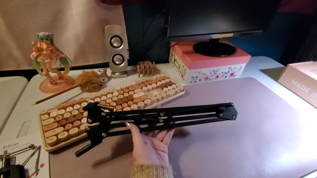 a bare, but cozy looking desk. an anime figurine, cute pen, flowered box and aesthetic keyboard are visible in the background.  heyshadylady is holding a boom arm for a microphone up with one hand. the folded up boom arm is about the same size as the keyboard.