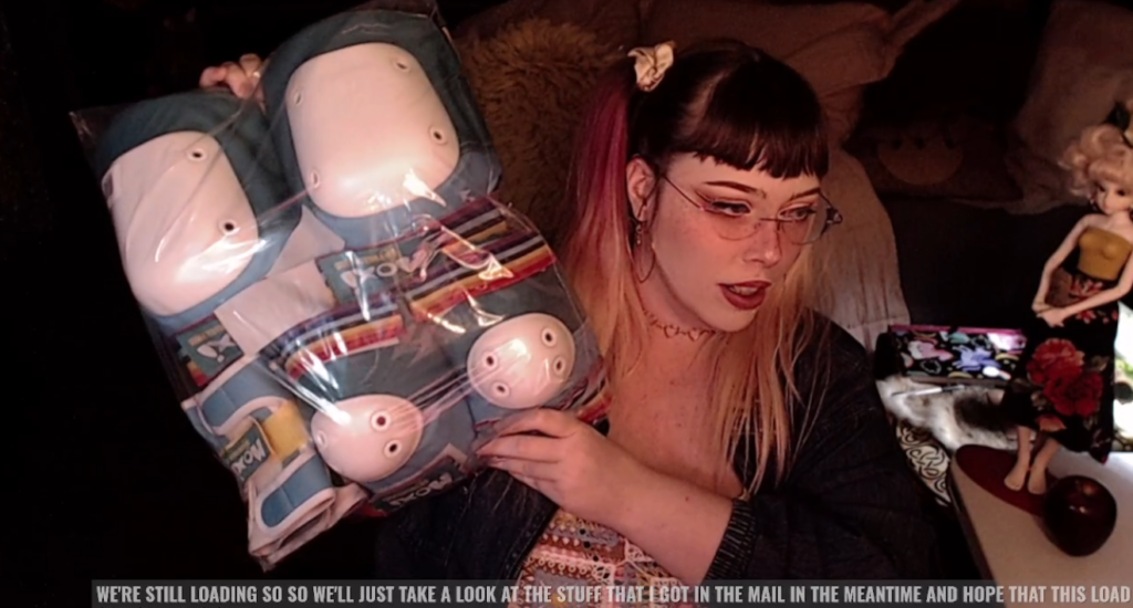 a screencap from my twitch livestream. my hair is up in pigtails, my makeup is done, and im holding up a wrapped package of kneepads, elbow pads and wrist guards. the caption at the bottom reads: "we're still loading, so we'll just take a look at the stuff i got in the mail in the meantime"