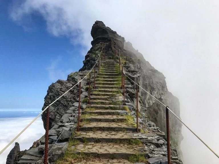 A picture of a tall jagged, pointy mountain surrounded by blue sky and fluffy clouds. There is a staircase carved into the mountain. The photo is shot as if you are standing at the base stair looking up at the journey ahead. You can see the stairs become smaller and smaller they further away they are as they reach the summit until eventually they disappear into the misty mountain top. There's railing around the stairs, but the journey seems long and a little treacherous.